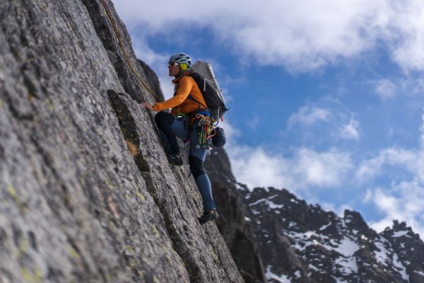 Horolezkyně s helmou a batohem leze po skalní stěně v mikině Rab Ascendor Hoody.